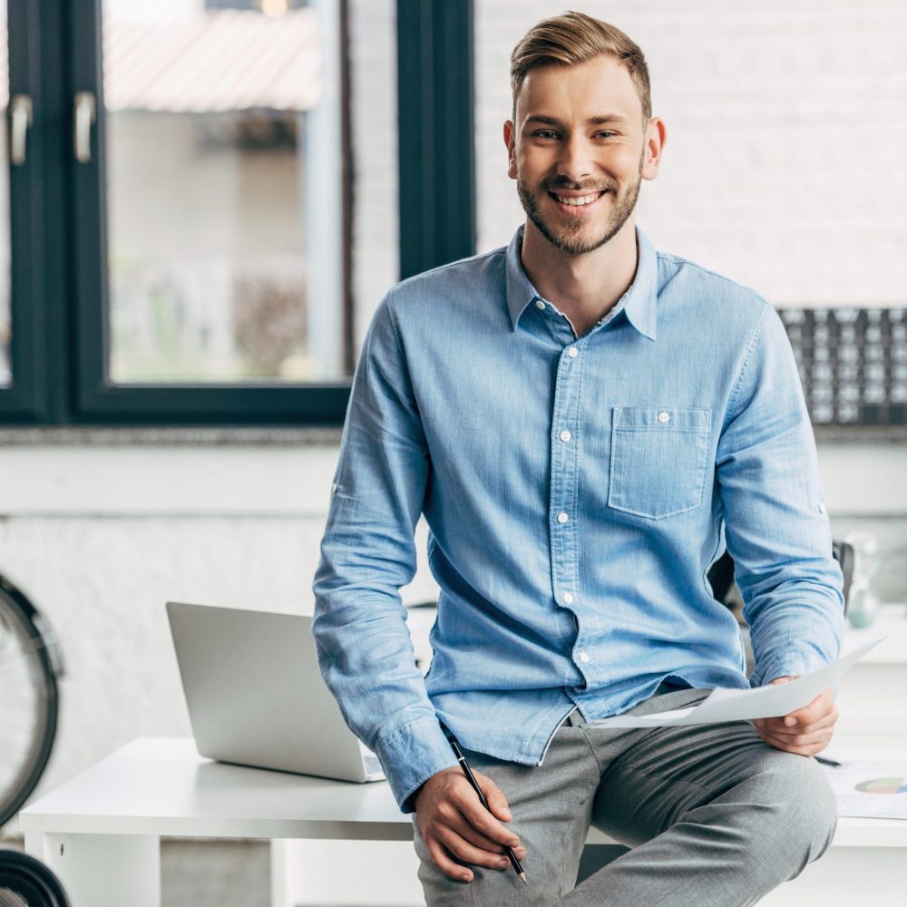 handsome young businessman holding papers and smiling at camera in office