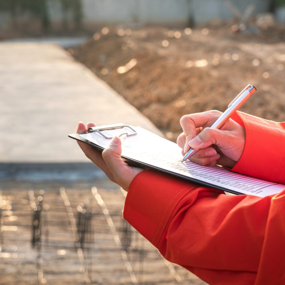 Action of a civil engineer is checking the building quality report form with construction work site as background. Industrial working action scene with people, close-up and selective focus.
